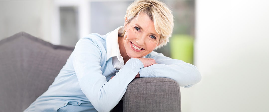 A mature woman leaning on the corner of a couch smiling.