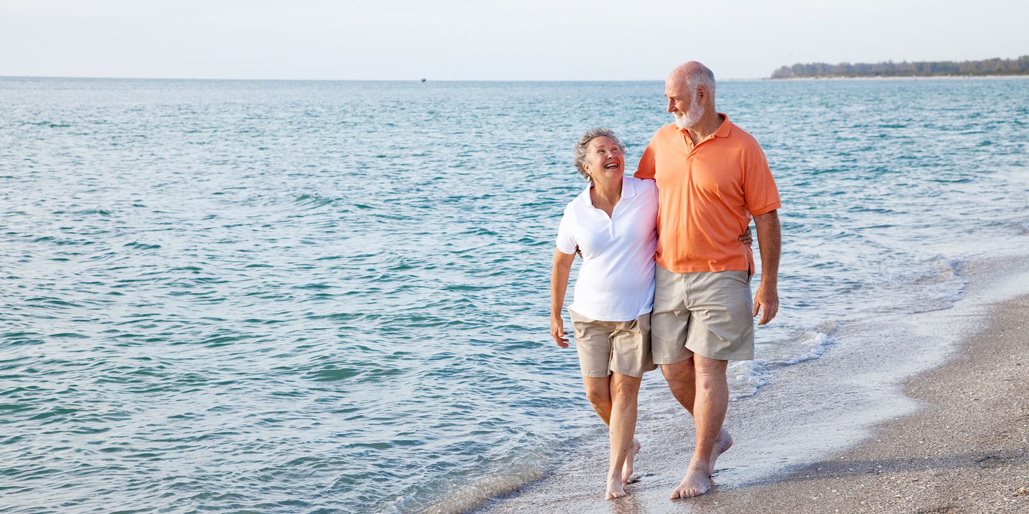 A senior couple enjoying coastal living as they walk barefoot along the beach shore, smiling together. 
